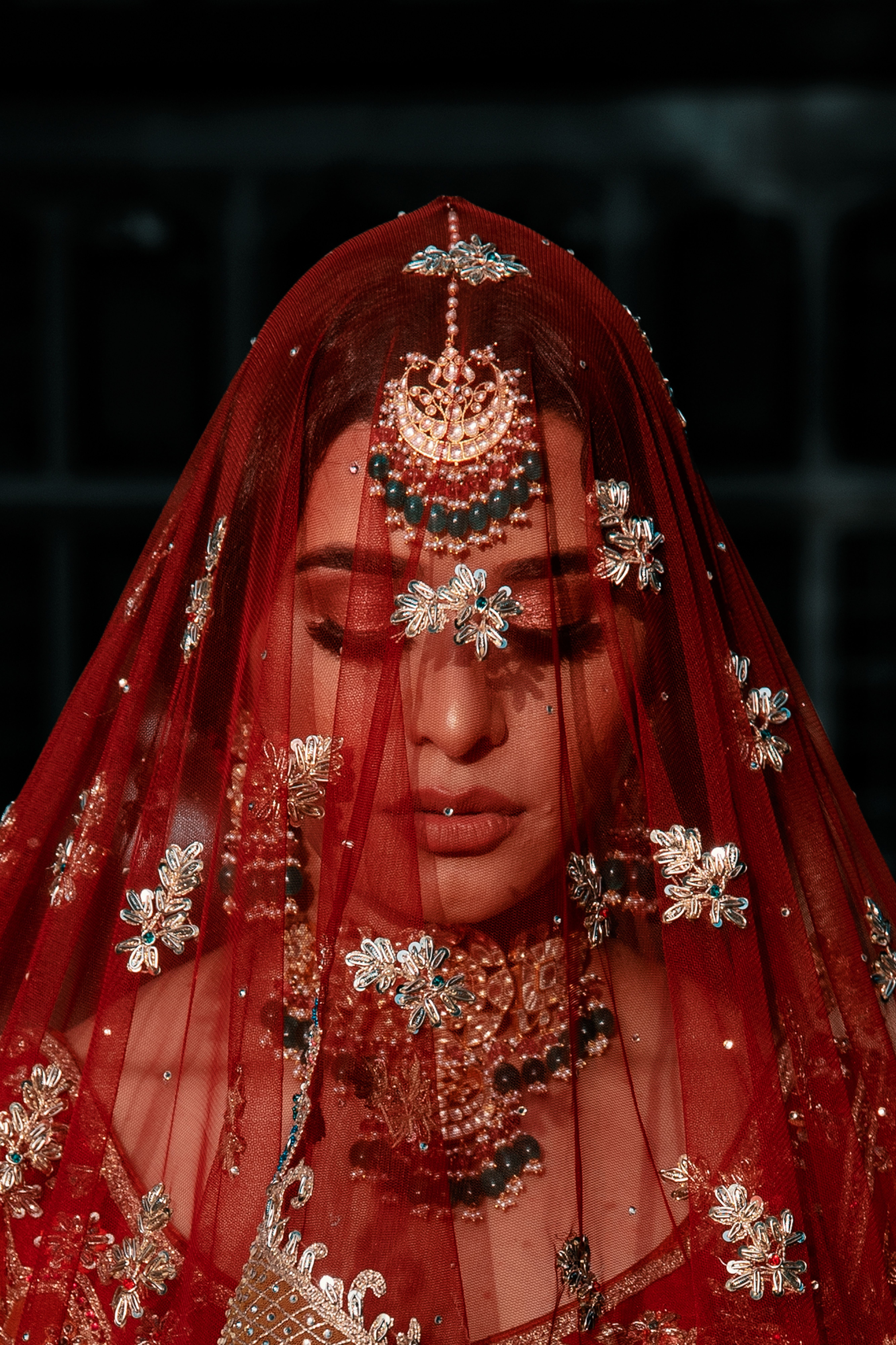 A bride's face resting beneath an embroidered red dupatta, eyes closed, her maang tikka shining through the veil — an inspiration photograph